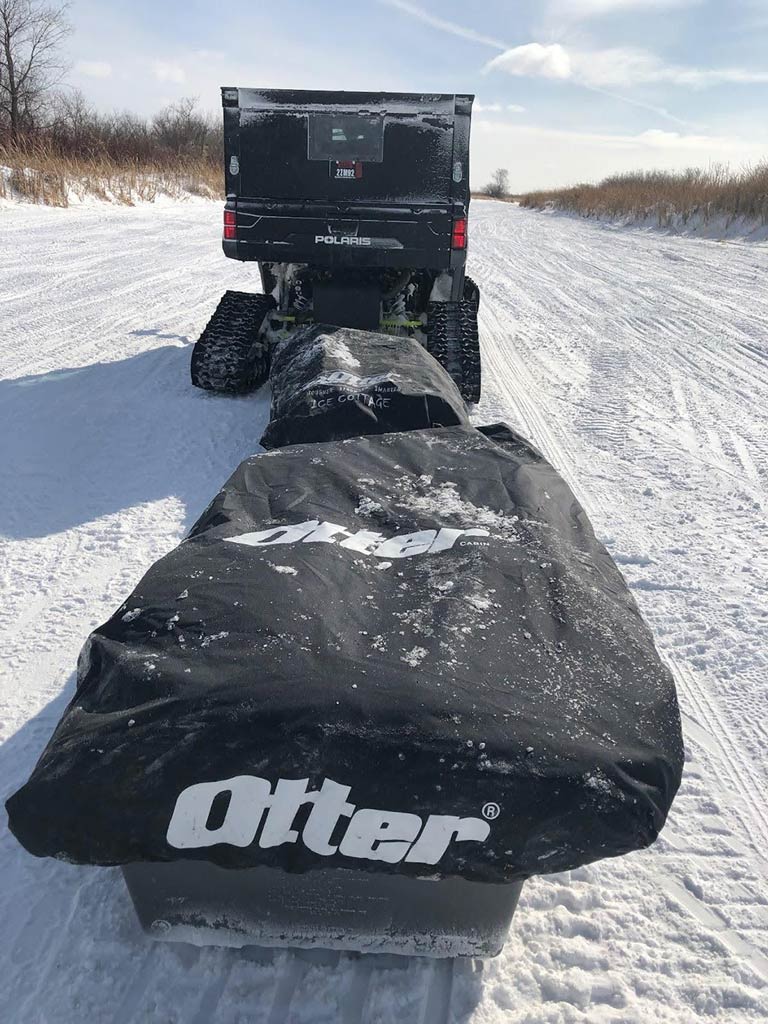 A snowmobile with tank-like tracks is towing a covered Otter ice fishing tent along a snow-covered trail, and dry grasses line the sides.