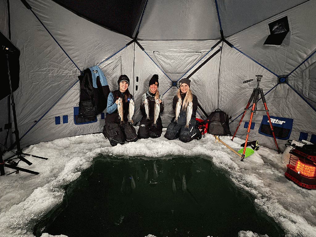 Three women kneel inside an ice fishing tent, each holding a couple of fish above a large hole in the ic, with fishing gear and a portable heater visible.