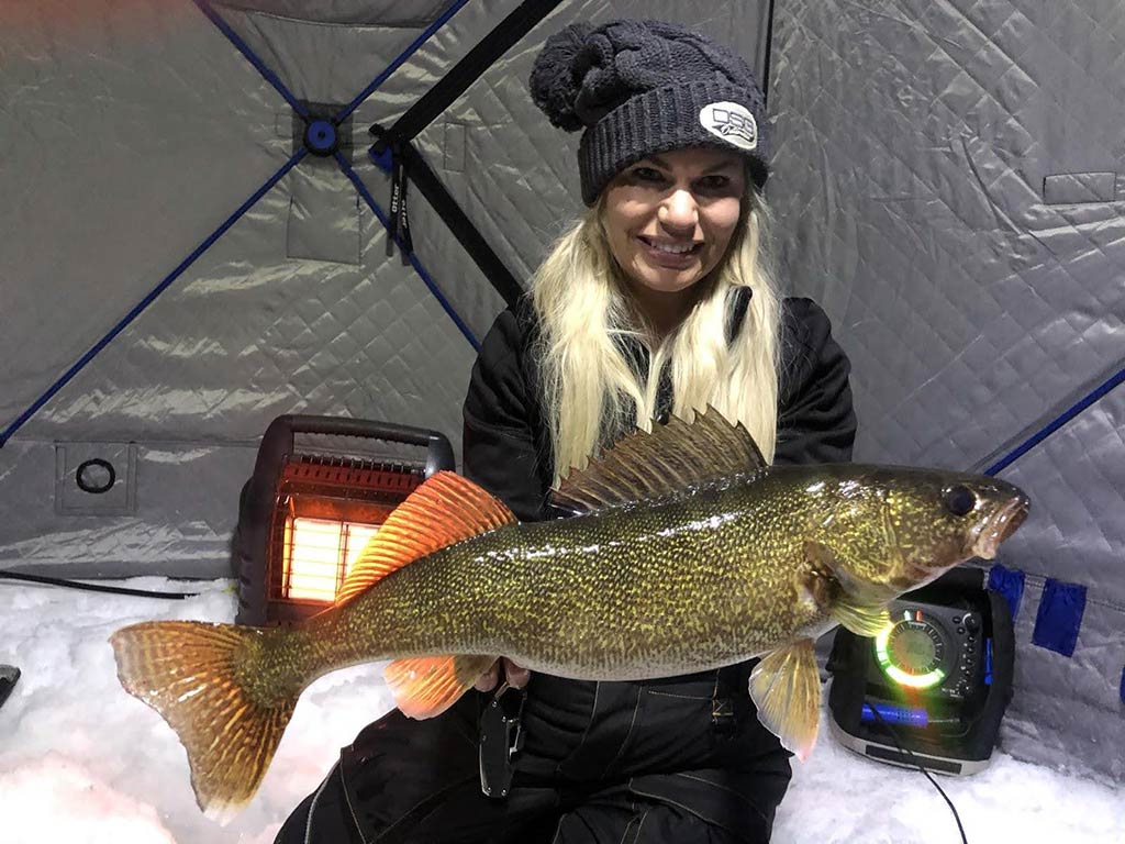 A woman in winter clothing holding a large Walleye fish inside an ice fishing shelter, with heaters and equipment visible in the background.