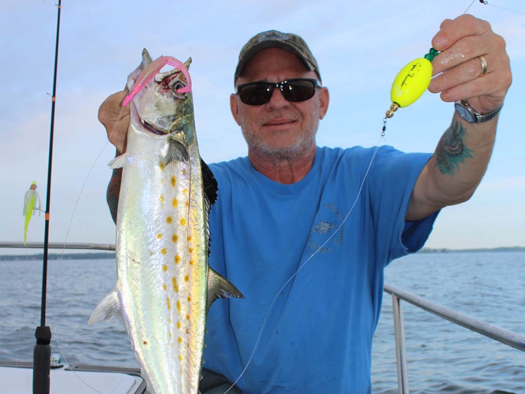 Мужчина в бейсболке и солнцезащитных очках показывает поппинг-корк и испанского макреля на лодке после удачного дня.