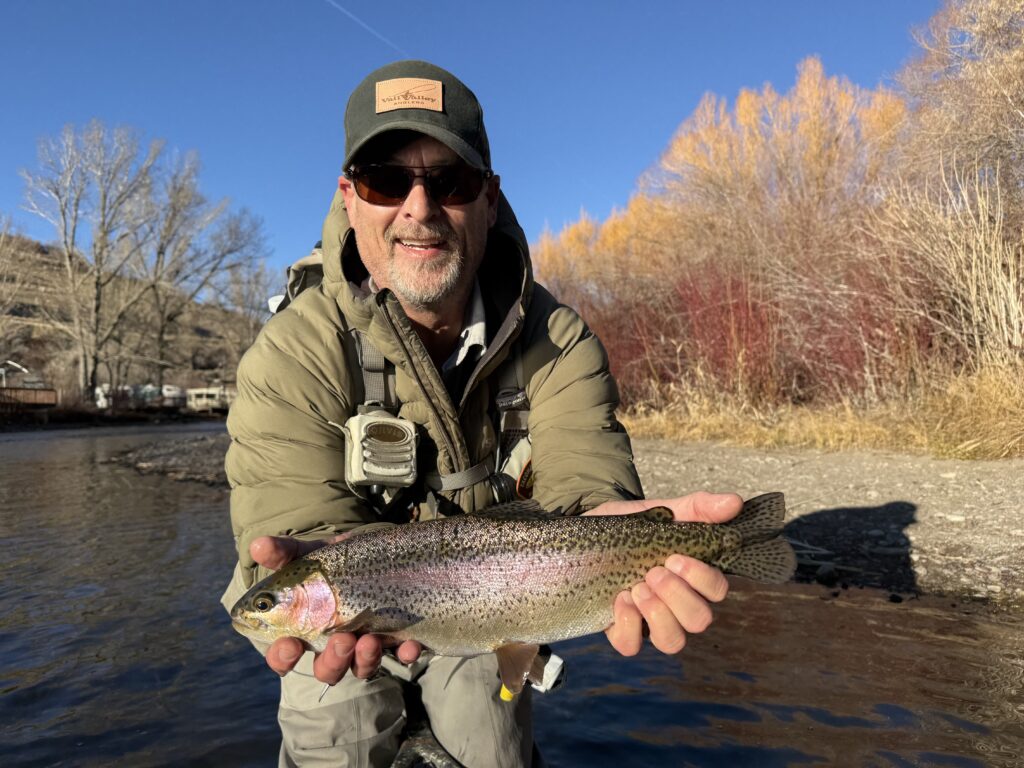 A person in outdoor gear's holding a large Rainbow Trout by a riverbank, with bare trees and autumn foliage in the background under a clear blue sky.
