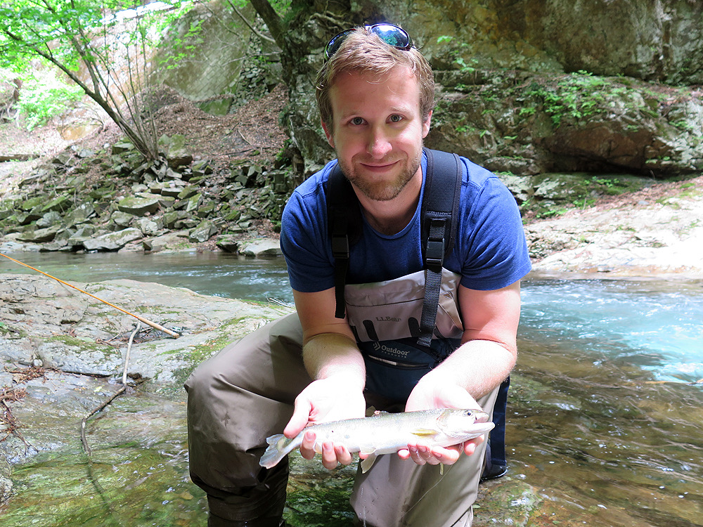 Tad Murdock in waders kneels by a rocky stream in a forested area, proudly holding a small trout.