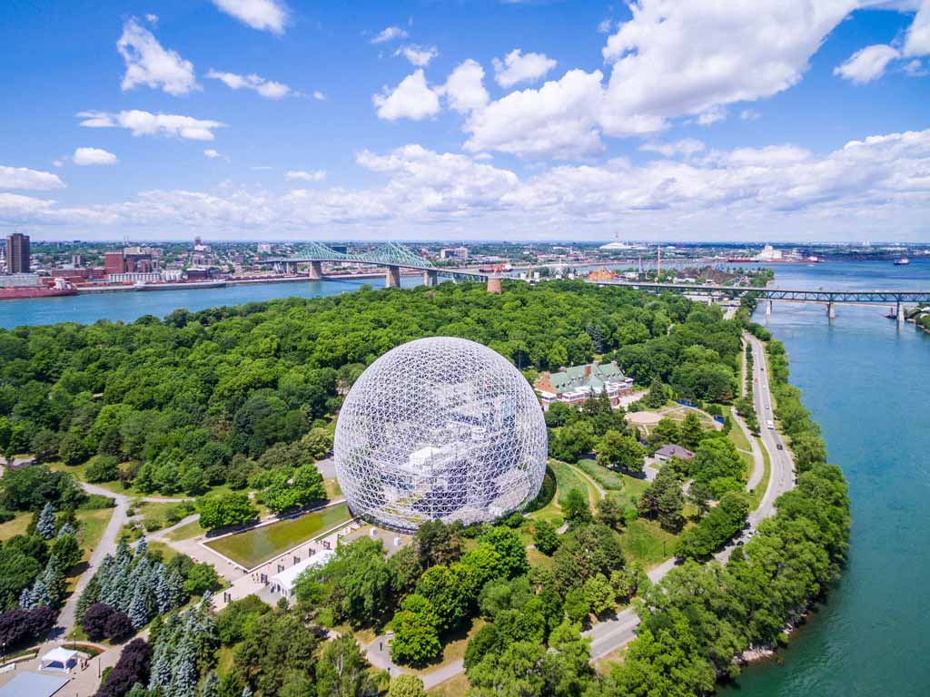 An aerial view shows a large geodesic dome surrounded by trees near a river, with a city and bridge visible in the background under a partly cloudy sky.