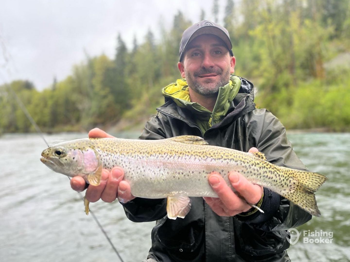 A man in outdoor gear holding a large Rainbow Trout by a river, showing why this spot is one of the best fishing cities in Canada, with trees beautifully framing the scenic background.