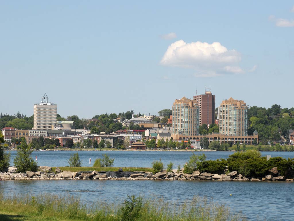 A skyline view of Barrie, Ontario, with some waters and greenery in the foreground and the city buildings in the distance.