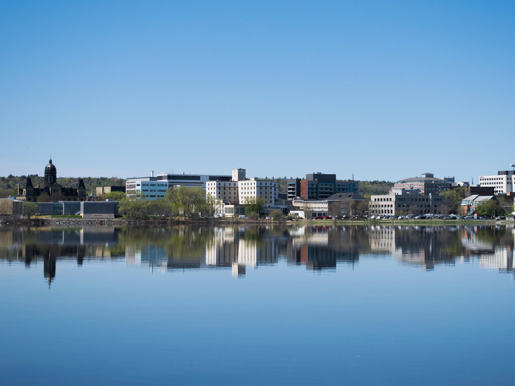 A view of Fredericton, NB from the water on a sunny day with clear skies. The city buildings are reflecting off the river surface.