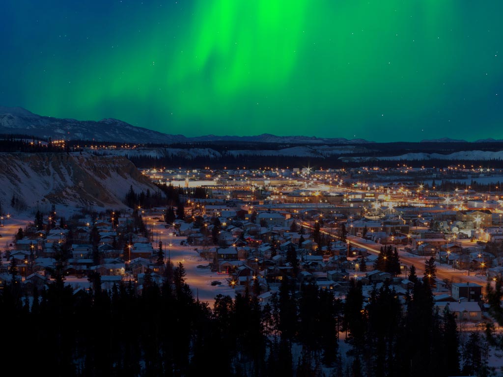 Northern lights light up the night sky above the snowy city of Whitehorse, one of Canada's best spots for fishing, with glowing buildings and trees all around.