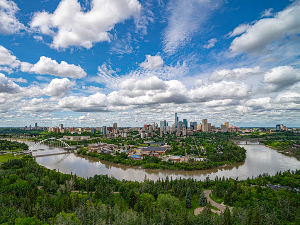 A view of the Edmonton skyline with a river winding in the forefront and lush greenery surrounding it.
