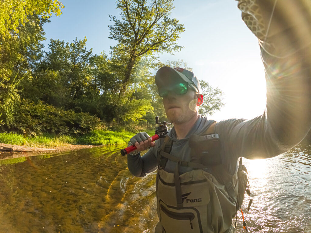 AJ Houser fishing in a river, possibly hoping to catch Pike or Bass. He&rsquo;s standing near the water, holding a rod, with trees lining the riverbank.