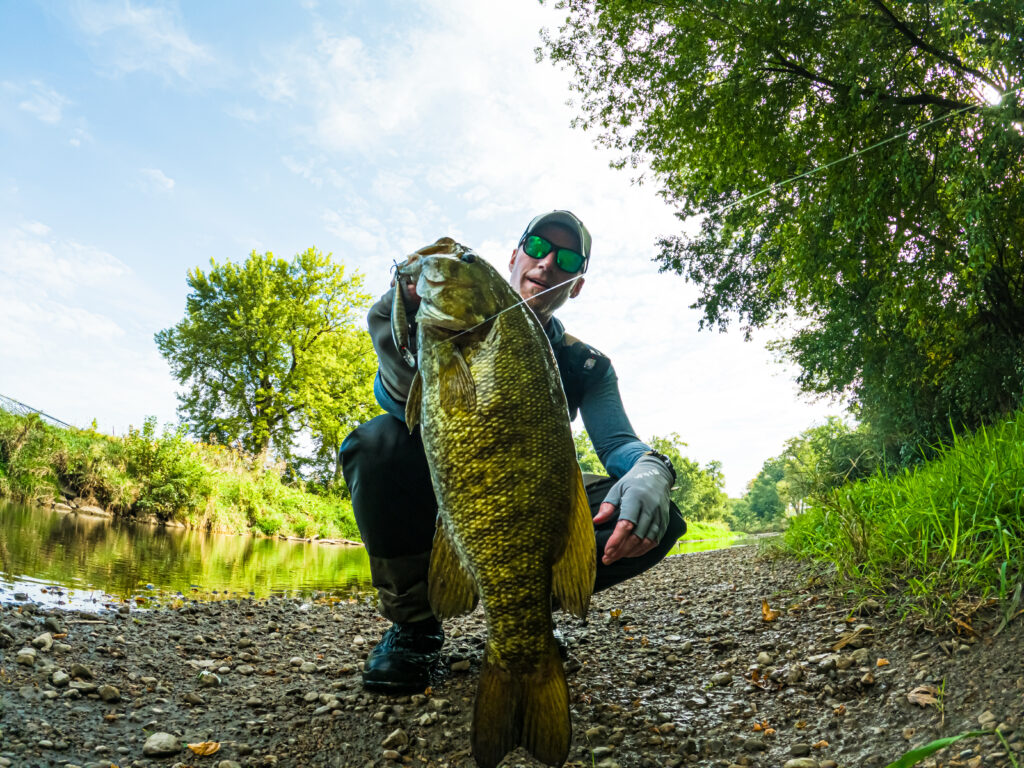 AJ Houser wearing sunglasses and gloves as he kneels on a riverbank, holding a large Bass with one hand. Trees and water are in the background.