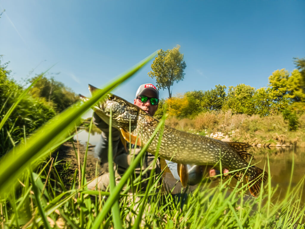 AJ Houser wearing sunglasses and a cap while holding a big Pike near a grassy riverbank, with trees and a blue sky behind them.