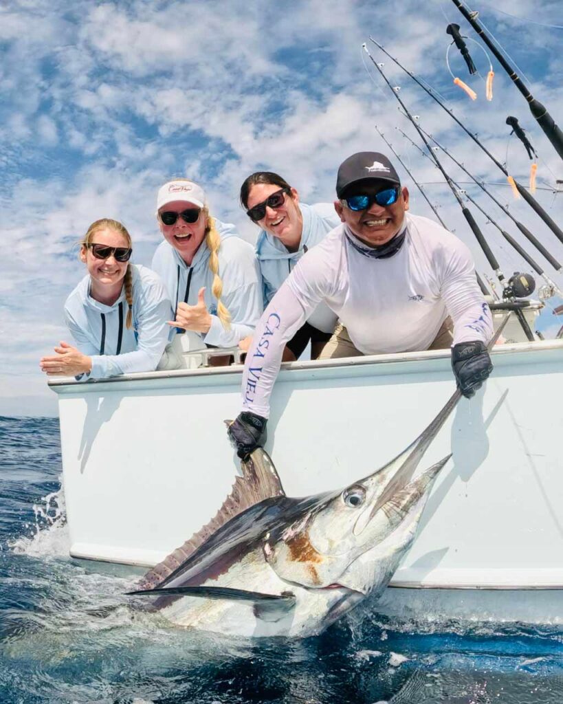 Becca Salmon, along with two of her IWFA friends and the boat captain leaning over the side of the boat, posing for a photo with a huge marlin that the captain is holding halfway out of the water.