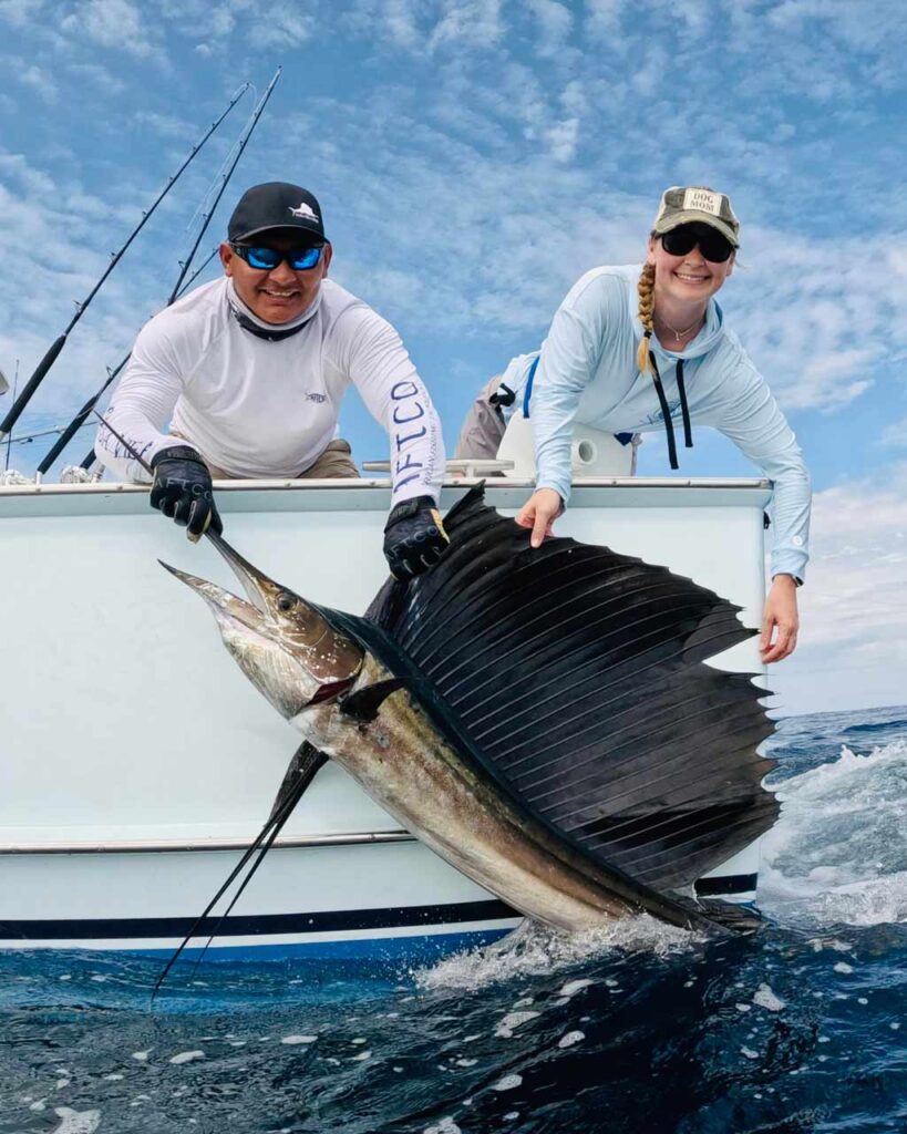 Becca Salmon leaning over the side of the boat and holding a Sailfish by its sail with the captain of the boat next to her holding the fish by its bill.