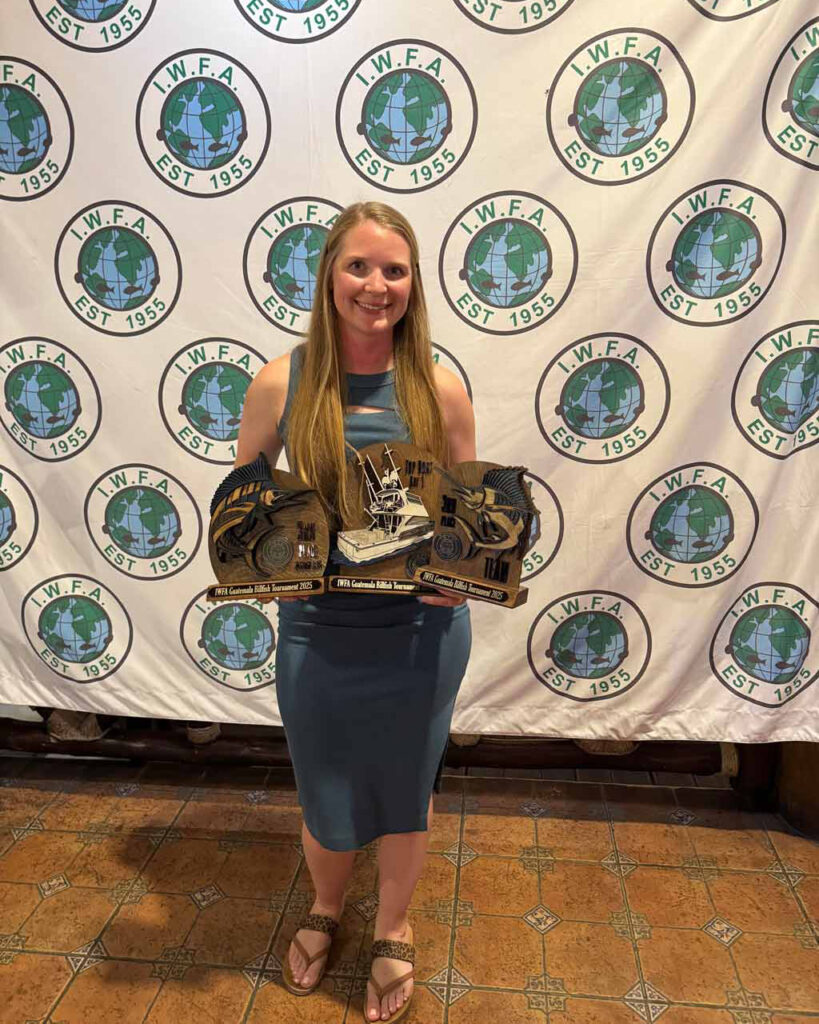 Becca Salmon in a blue dress, standing indoors, holding three Casa Vieja tournament trophies in front of an IWFA backdrop.