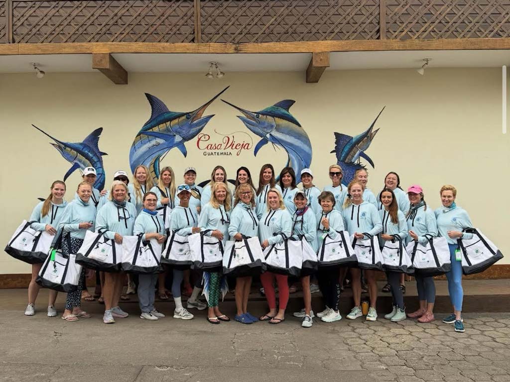 IWFA women in matching light blue hoodies pose with bags in front of a mural featuring Marlin and the words "Casa Vieja Guatemala" on the wall.