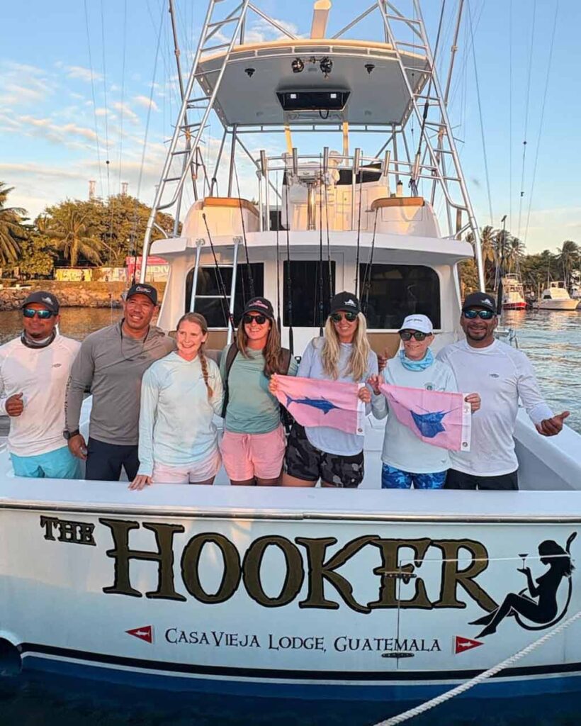 Four IWFA members are standing on a docked fishing boat called "The Hooker" at Casa Vieja Lodge, Guatemala, holding flags with a Fish illustration, with three boat crew members besides them.