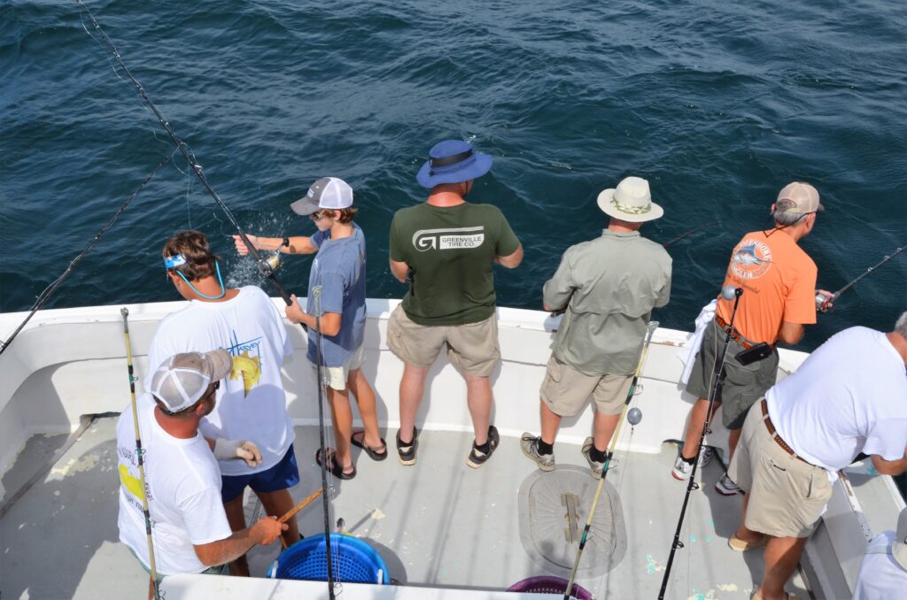 Six people are standing at the edge of a boat, fishing over the side with rods angled out over the water. The view’s from above.