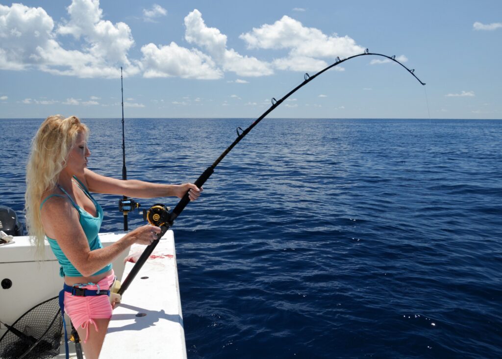 A woman on a boat holds and bends a fishing rod over deep blue ocean water under a partly cloudy sky.