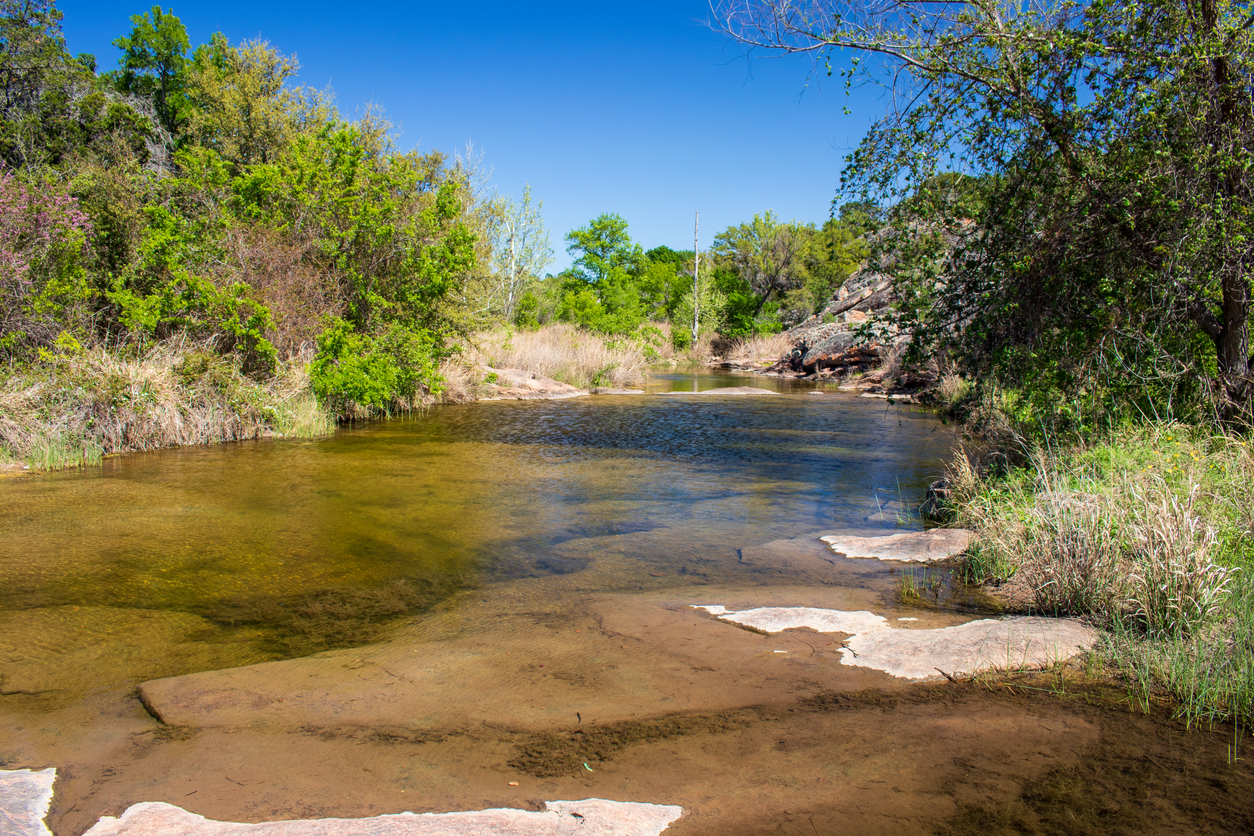 A shallow creek with clear water flows through one of the Top Ten Fishing State Parks in Texas, surrounded by rocks, grass, and dense green trees under a bright blue sky.