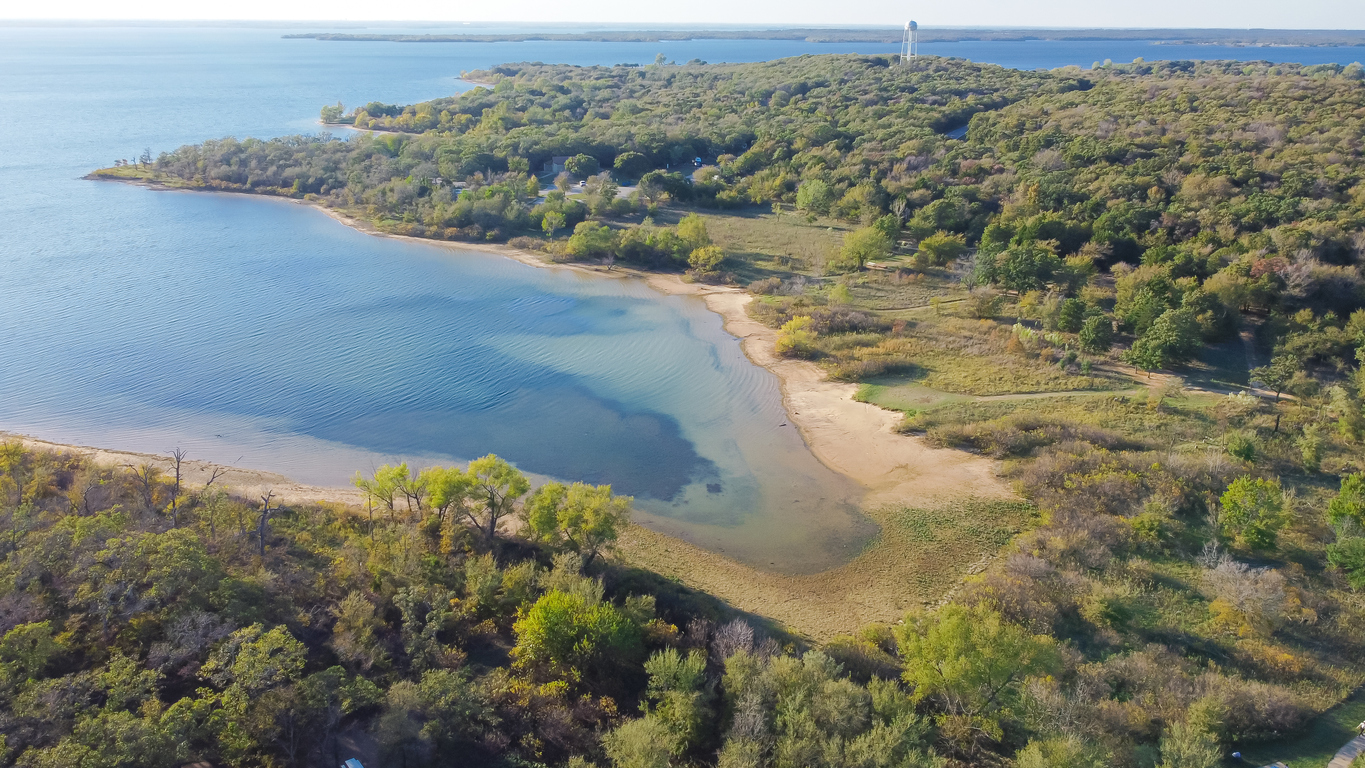 You’re seeing an aerial view of a small sandy beach and clear water surrounded by dense green trees at one of the top ten fishing state parks in Texas, along a scenic lakeshore.