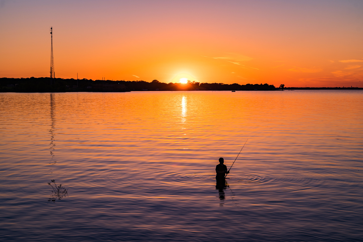 Someone's standing in calm water, fishing at sunset. The sky's orange, and a distant shoreline shows—it's one of the top ten Fishing State Parks in Texas.