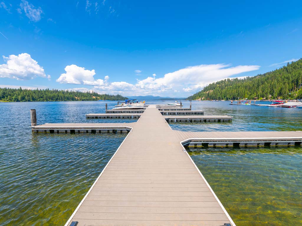 A shot of a wide wooden dock on Priest Lake, one of the best Trout lakes, with boats on both sides and forested hills beneath a sunny, cloud-filled sky.