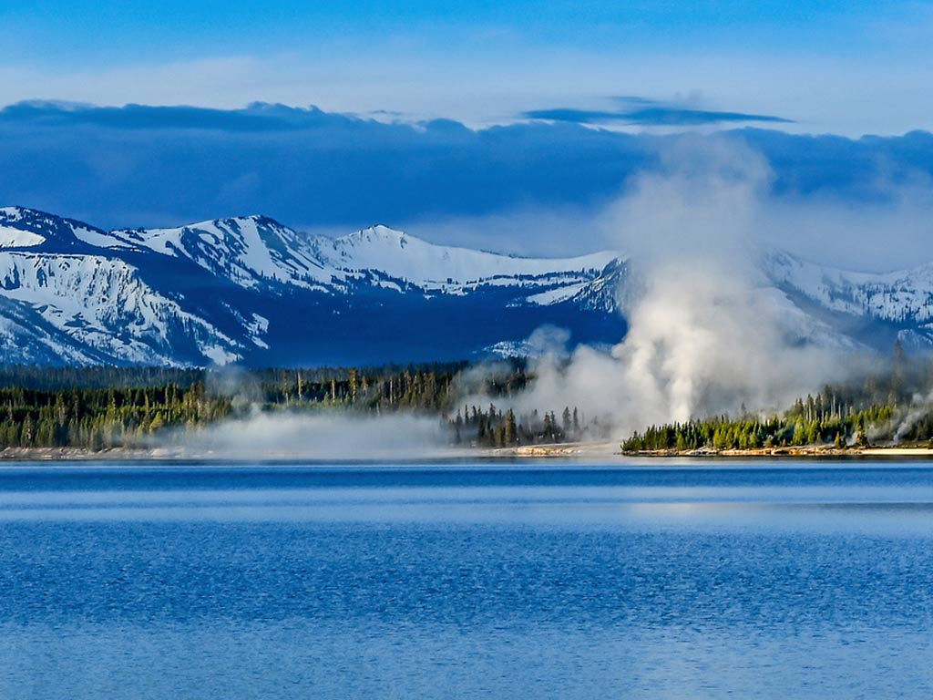 Yellowstone Lake with steam rising from geysers on the shore, evergreen trees, and snow-capped mountains under a partly cloudy sky.