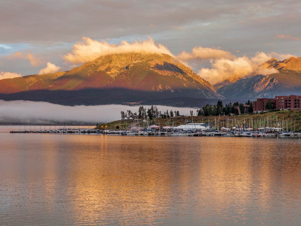 A shot of the marina on Lake Dillon with lots of docked boats, with mountains and some buildings behind it under a partly cloudy sunset sky.
