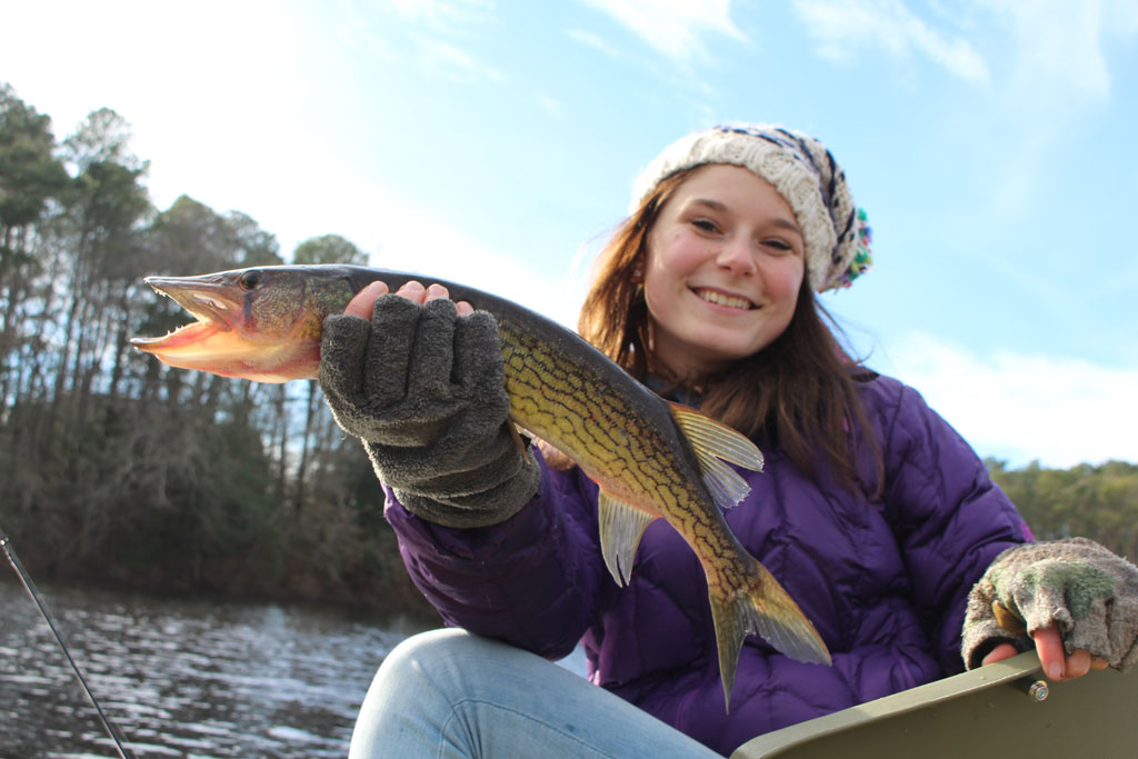 Someone in a purple jacket and knit hat's holding a large Fish while sitting in a boat on a lake, with trees and sky behind them.
