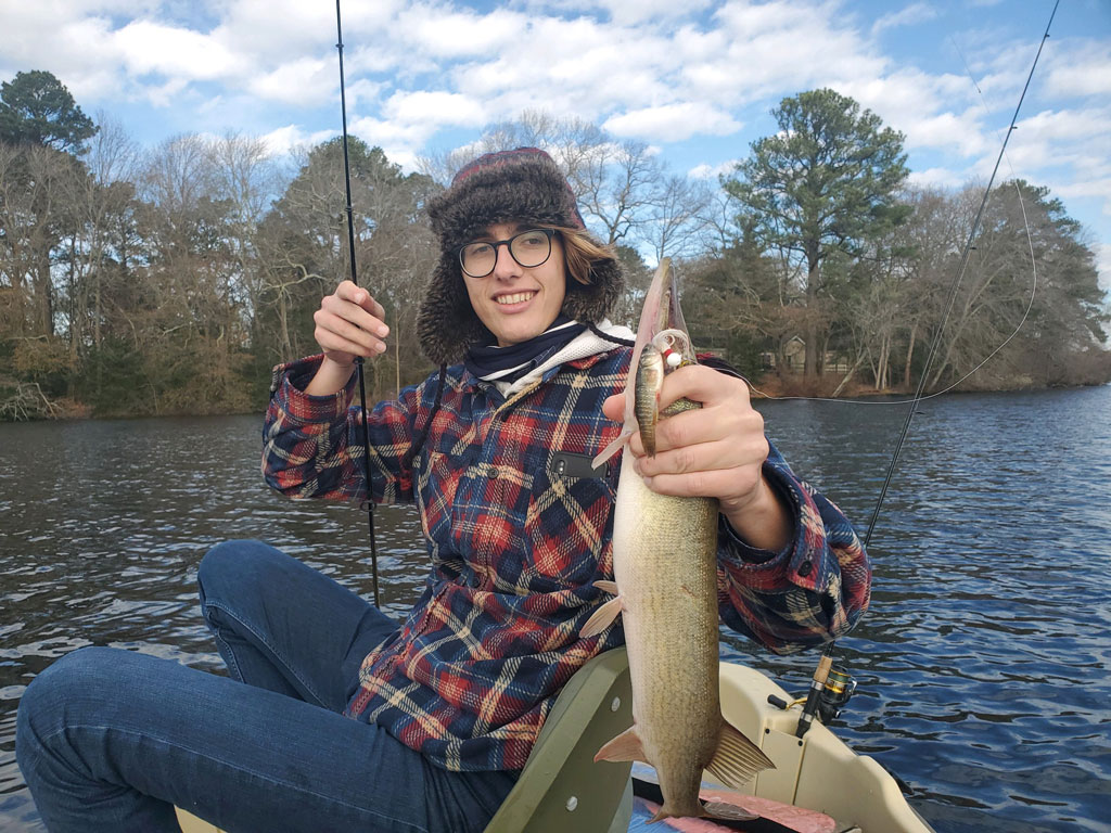 Someone's sitting in a boat on a lake, wearing glasses and a plaid jacket, holding up a freshly caught Trout with trees and water behind them.