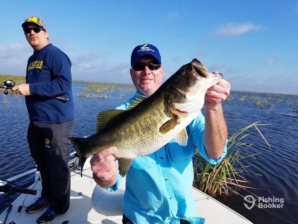Two men are on a boat; one’s holding a large Florida Bass and smiling at the camera, while the other’s fishing in the background on Okeechobee lake.