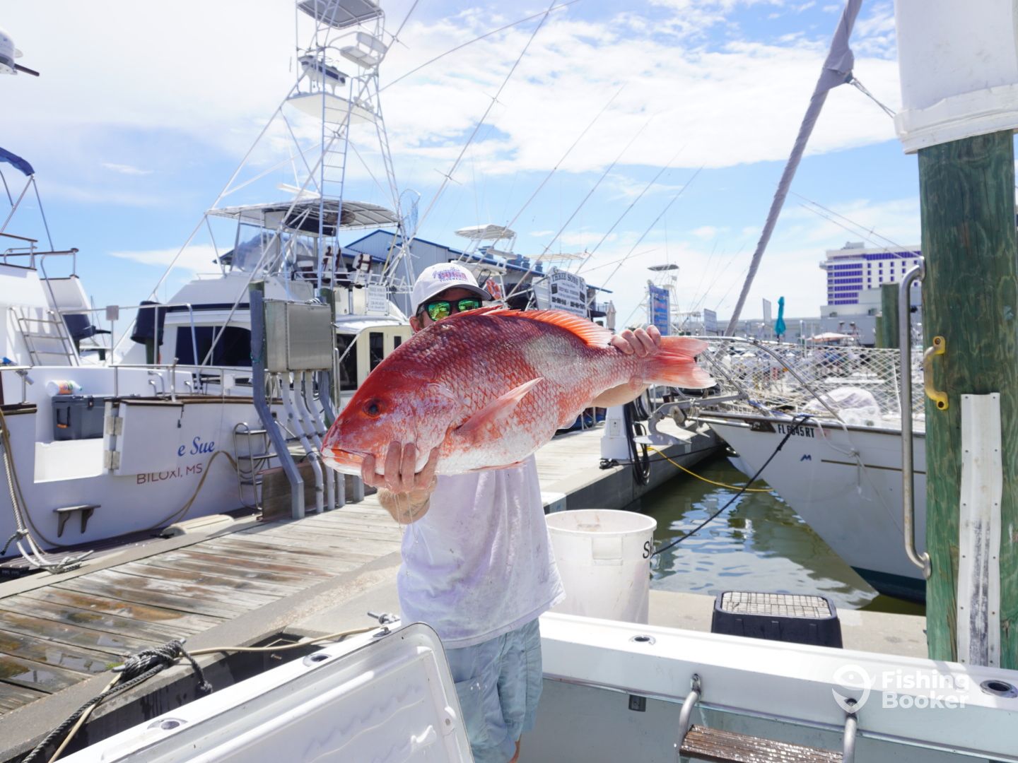 Person holding a large Red Snapper on a dock, with several boats in the background and clear skies above.