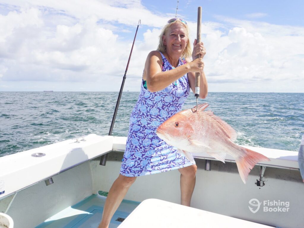 A woman in a blue patterned dress is holding a Red Snapper on a fishing boat in open water under a partly cloudy sky.