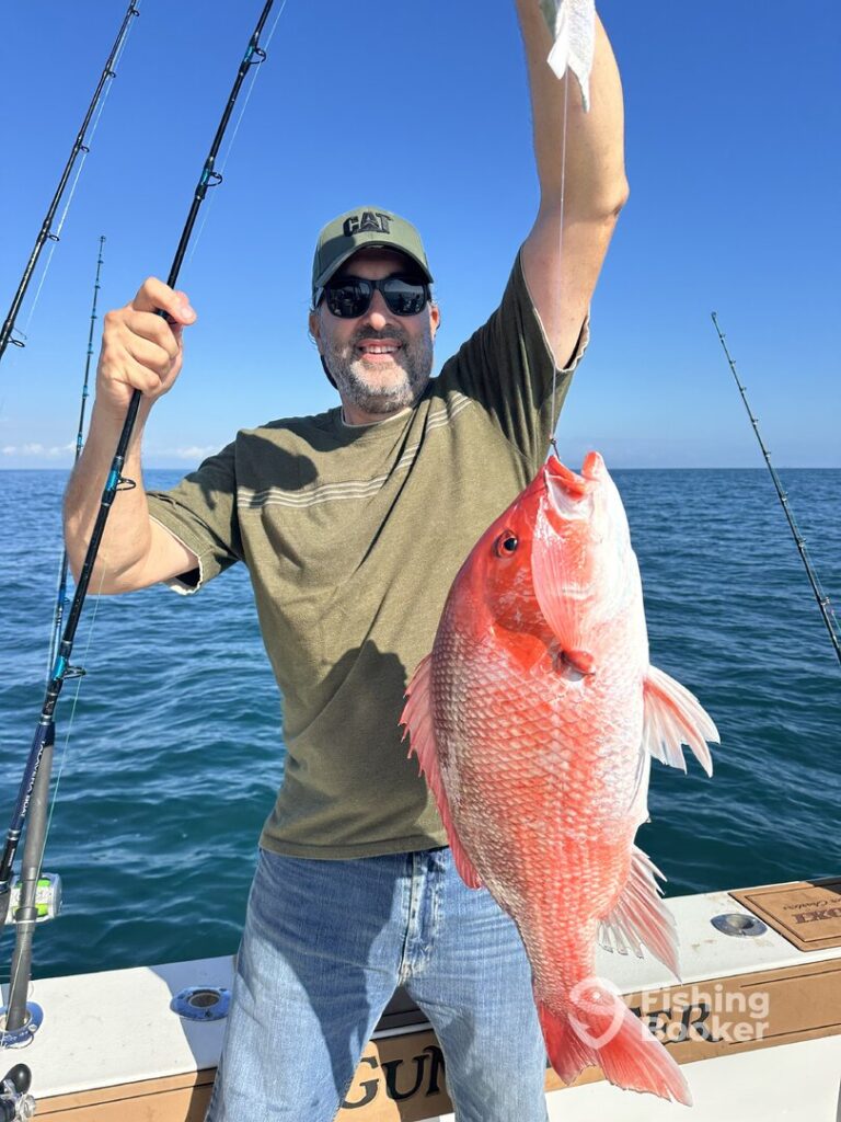 Man's wearing sunglasses and a cap, holding up a large Red Snapper fish on a boat with fishing rods; the ocean and sky are visible in the background.