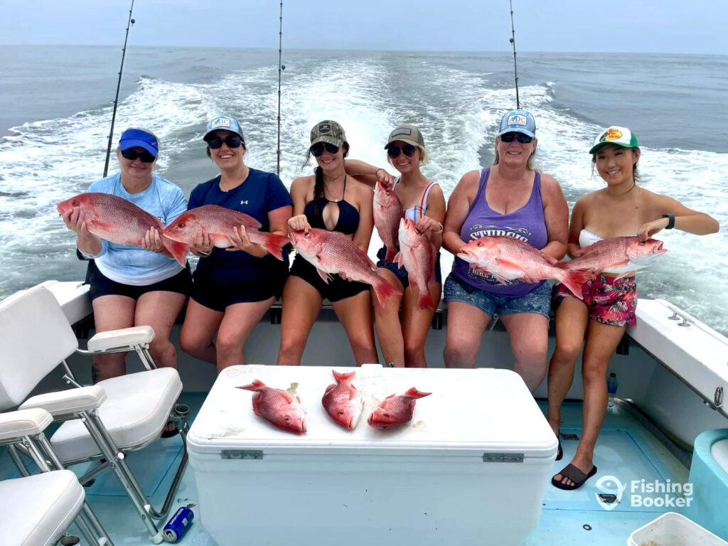 Six people on a fishing boat hold up Red Snapper they’ve caught, with more Red Snapper displayed on a cooler. The sea and fishing rods are in the background.