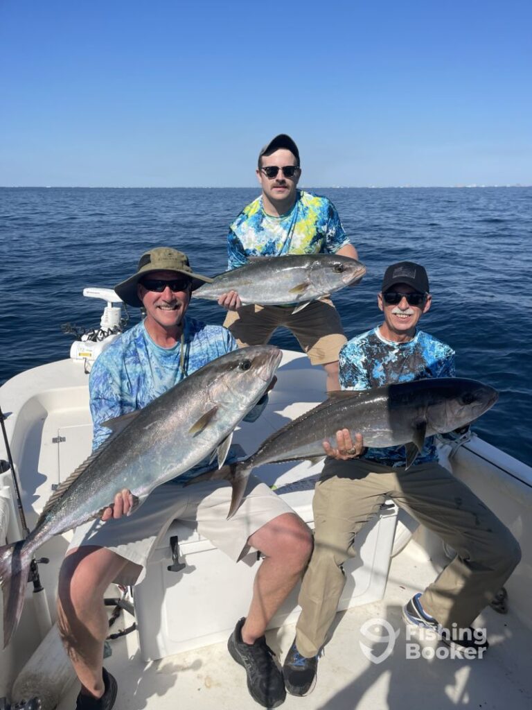 Three men are sitting on a boat during Amberjack season, each holding a large Amberjack. They're wearing matching blue shirts, and the ocean's in the background.
