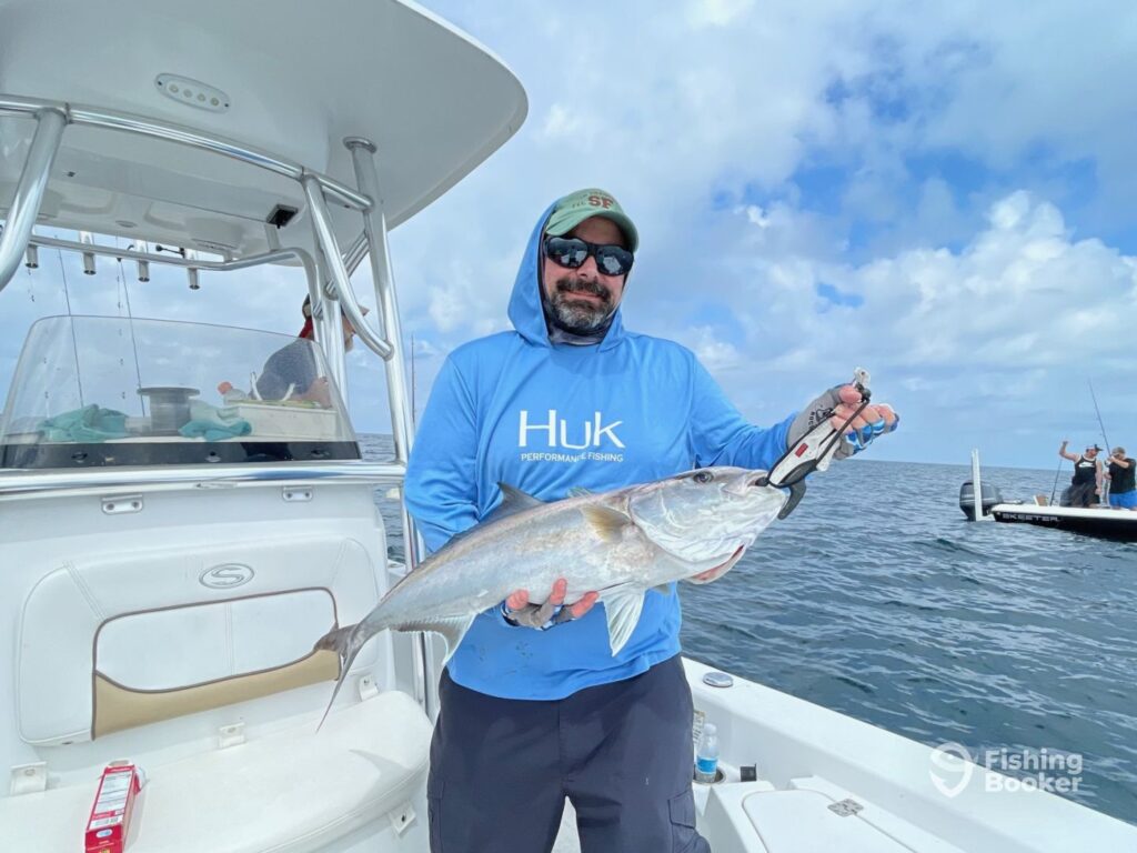 A man in a blue hoodie stands on a boat, holding a large Amberjack with pliers and enjoying the ocean view during Amberjack season. Another boat's visible in the background.