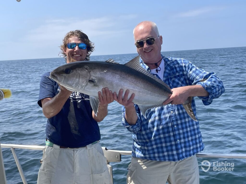 Two men on a boat are holding a large Amberjack, smiling brightly during Amberjack season, with the ocean and sky in the background.