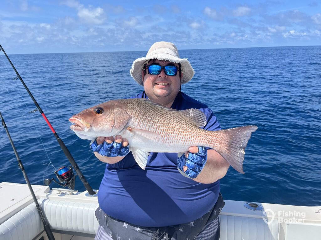 Someone on a boat holding a large Mangrove Snapper. Fishing rods stand behind him as the ocean stretches out under a partly cloudy sky.