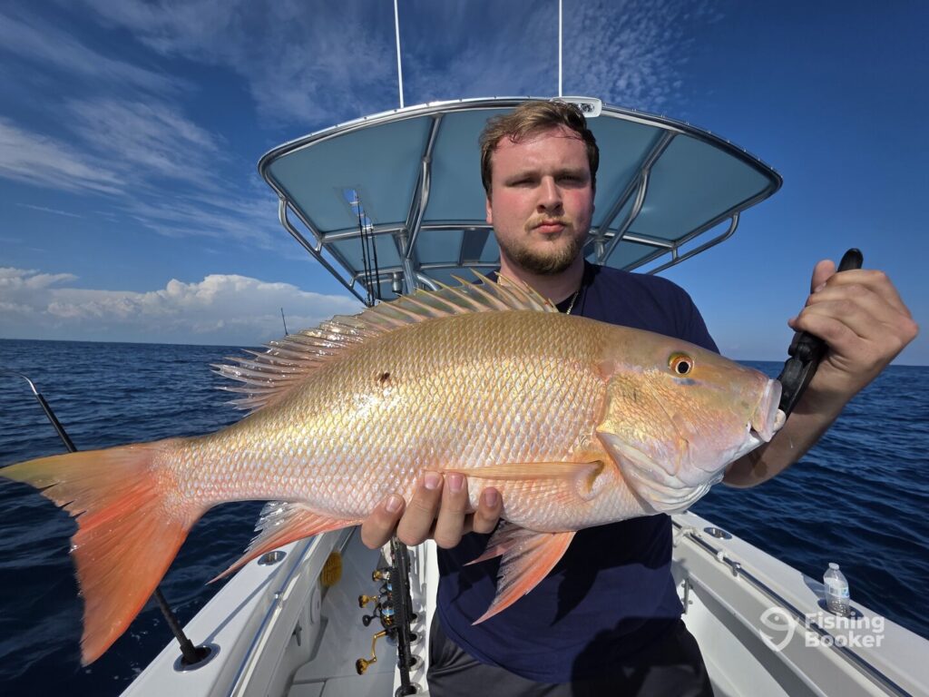 A man’s standing on a boat, holding a large Mutton Snapper, set against the open ocean and blue sky.