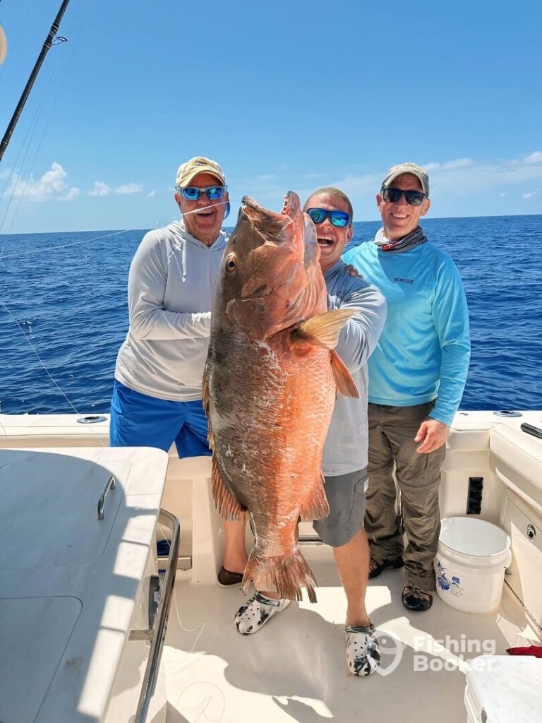 Three people on a boat hold a large Cubera Snapper, smiling at the camera during Snapper season in Florida. The ocean and blue sky are in the background.