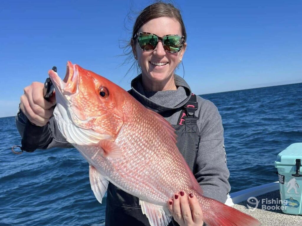 A woman wearing sunglasses holds up a large Red Snapper on a boat during snapper season in Florida, with the ocean in the background.