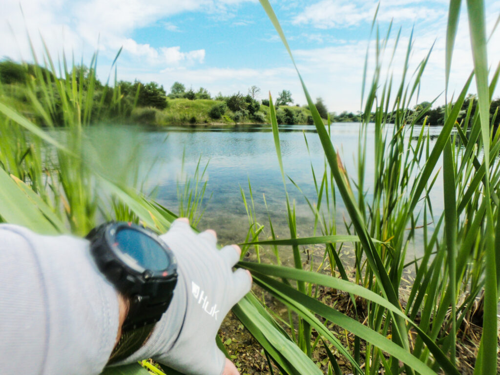 Someone's wearing a glove and wristwatch, holding tall grass near a clear, calm lake&mdash;it's the perfect place to learn how to catch big Bass with green trees and a blue sky all around.