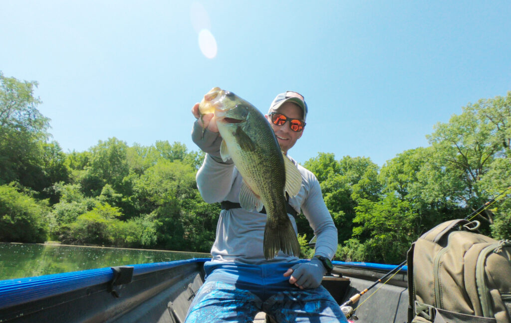 A person wearing sunglasses and a hat's sitting in a boat, holding up a large Bass to show how to catch big Bass, with fishing gear and green trees in the background.