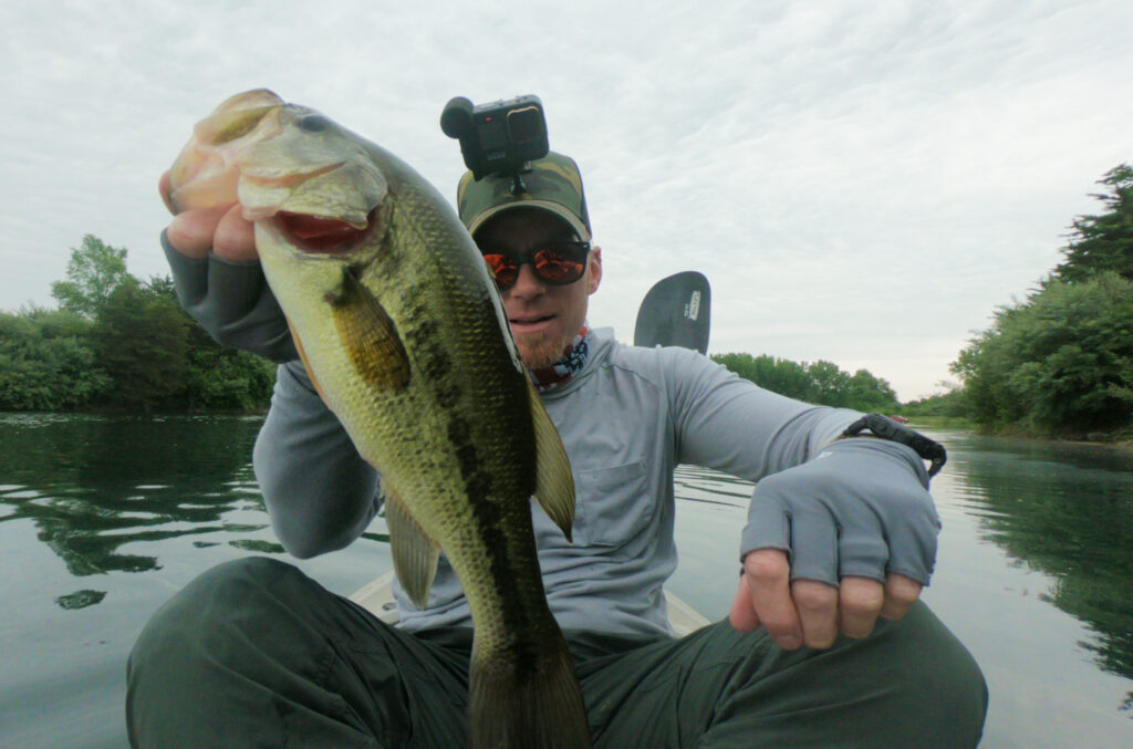 A person in outdoor clothing and sunglasses holds up a Largemouth Bass while sitting on a kayak in a calm, tree-lined lake, showing how to catch big bass. There's a camera on their head.