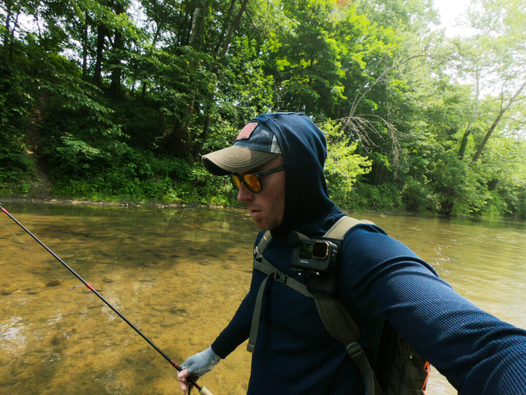 Someone's wearing outdoor gear and sunglasses while standing in a shallow, clear stream among green trees, showing how you catch big Bass with a fishing rod.