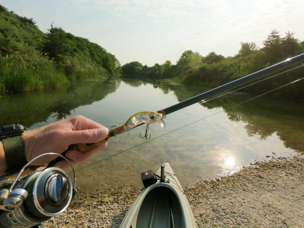 Someone's holding a fishing rod with a lure by a calm river, standing near a kayak&mdash;it's the perfect spot to learn how to catch big Bass, surrounded by trees and lush green foliage.