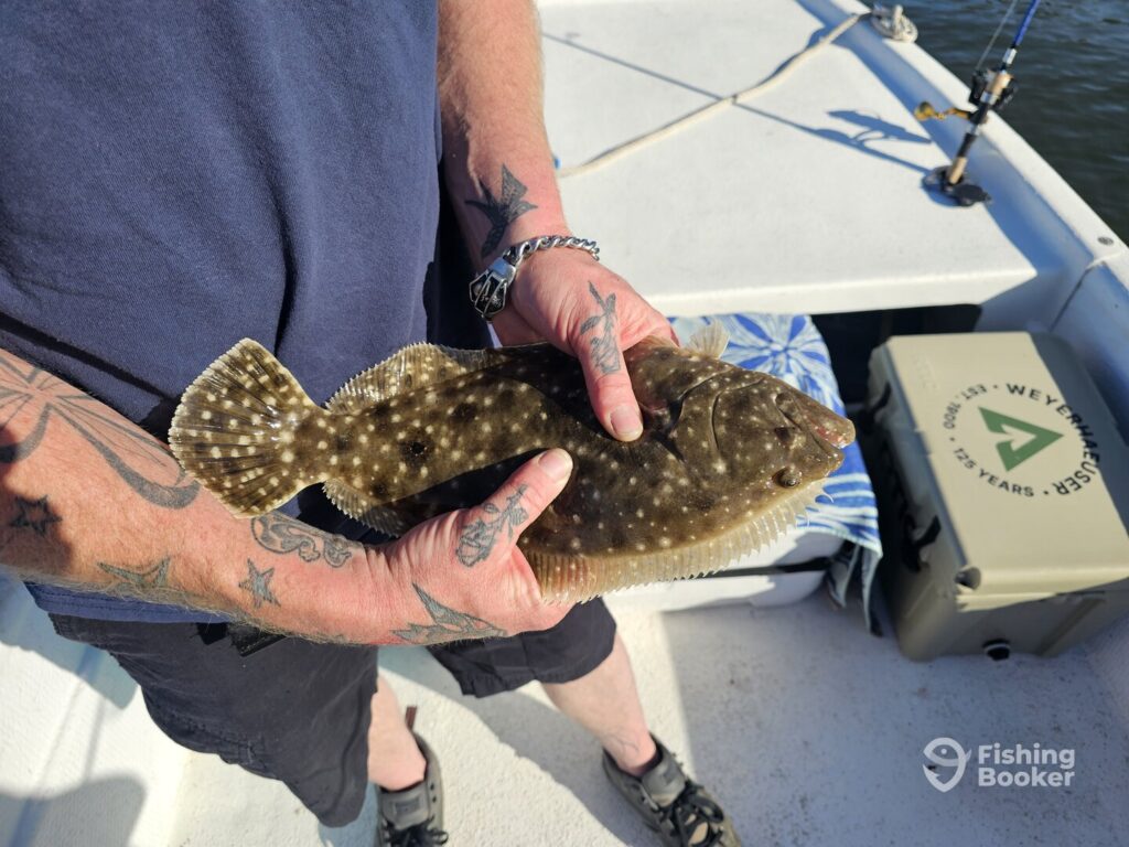 Person with tattoos is holding a Flounder fish on a boat, and there&rsquo;s fishing gear plus a cooler in the background.