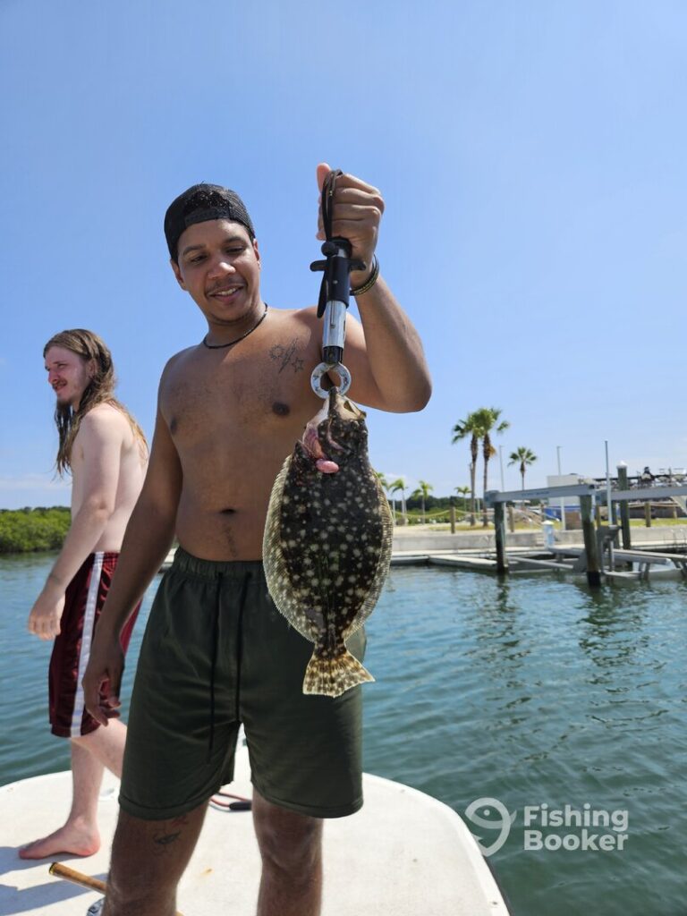 A man&rsquo;s standing on a dock, holding up a flat, spotted Flounder with a metal gripper; another person&rsquo;s nearby with water and palm trees in the background.
