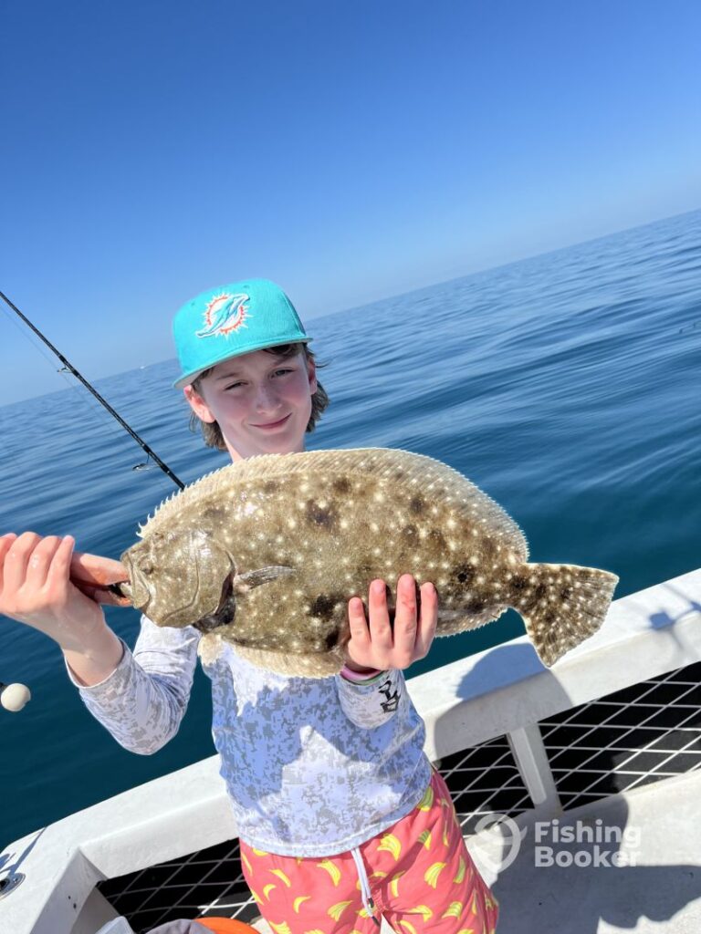 A person on a boat's holding a large Flounder, with calm ocean and blue sky behind. A fishing rod's visible; the person wears a turquoise cap and patterned shorts.